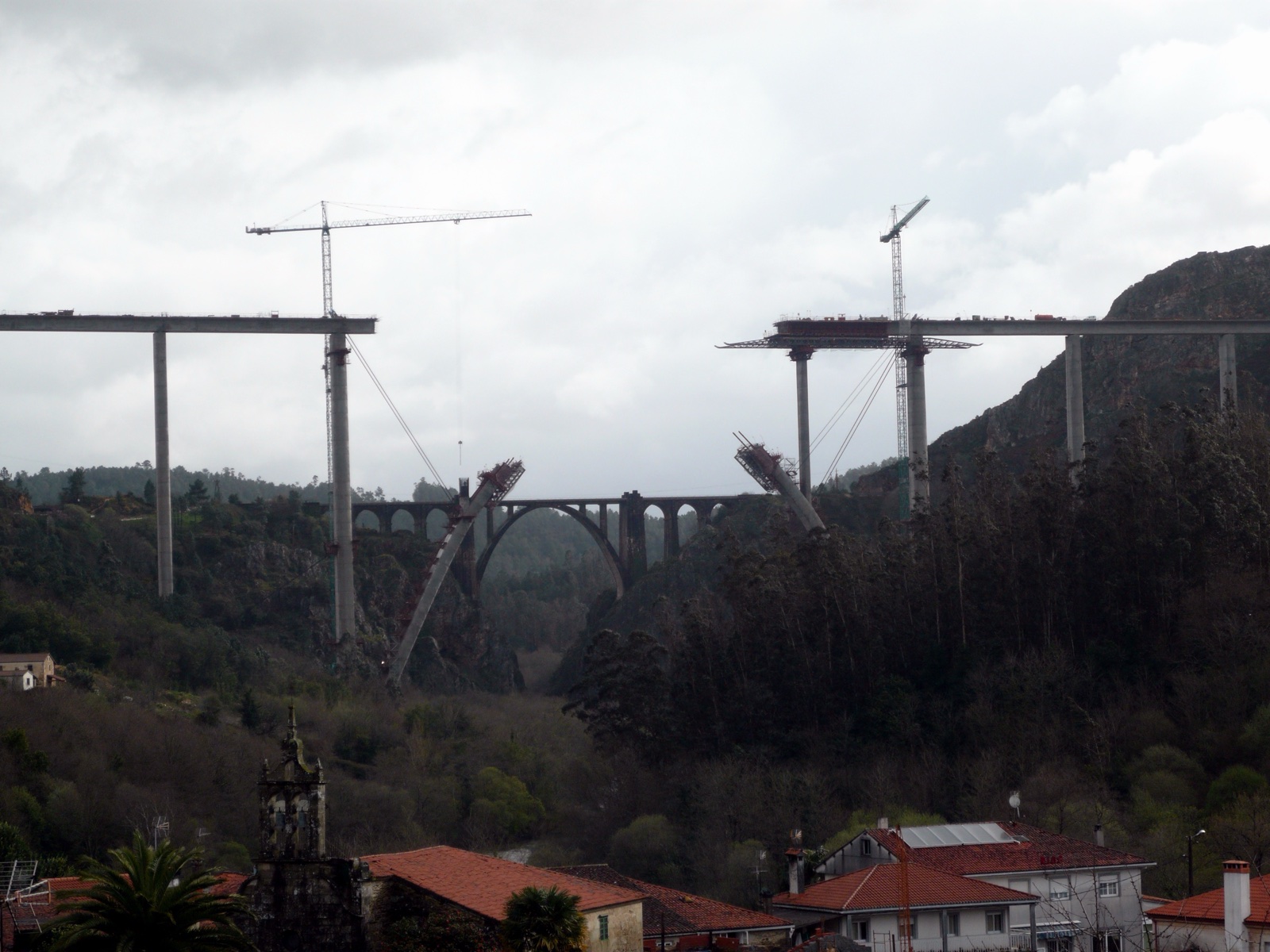 Dos tramos de un viaducto de hormigón en construcción, sobre pilares altos y con grúas, cruzan un valle boscoso por encima de un puente de piedra antiguo; abajo, tejados de un pueblo.