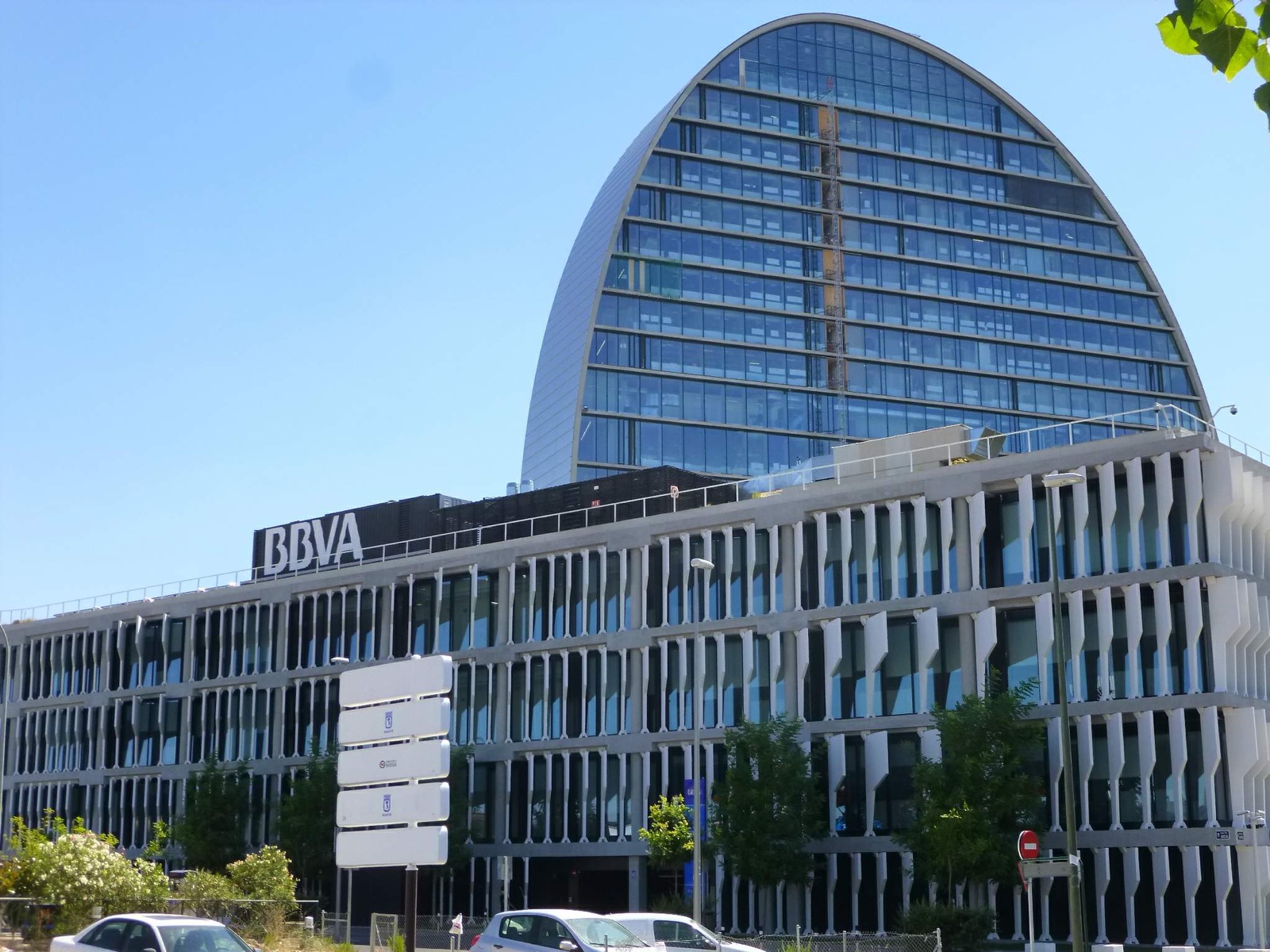 Edificio de oficinas bajo con el rótulo BBVA y, detrás, una torre de cristal con forma de vela, bajo un cielo azul.