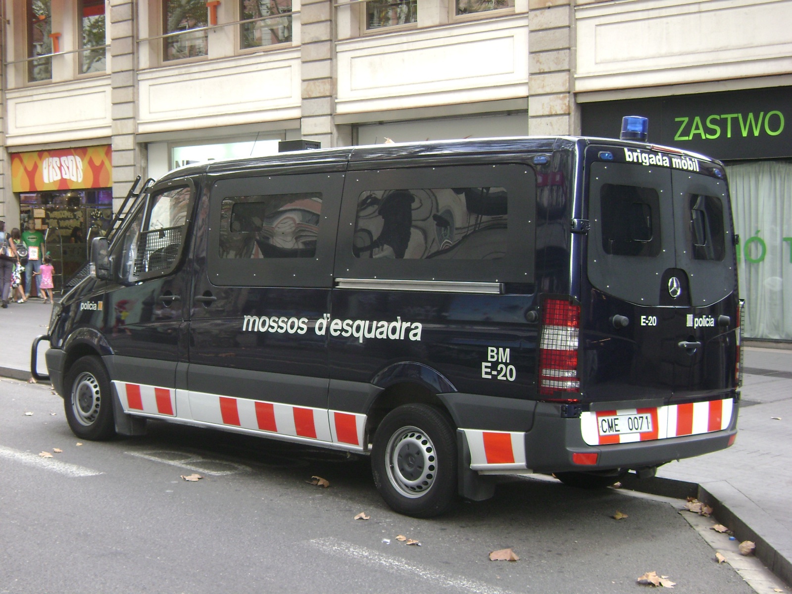 Furgoneta azul oscuro con el rótulo «mossos d'esquadra» y franjas rojas y blancas, aparcada junto a la acera ante unas tiendas.