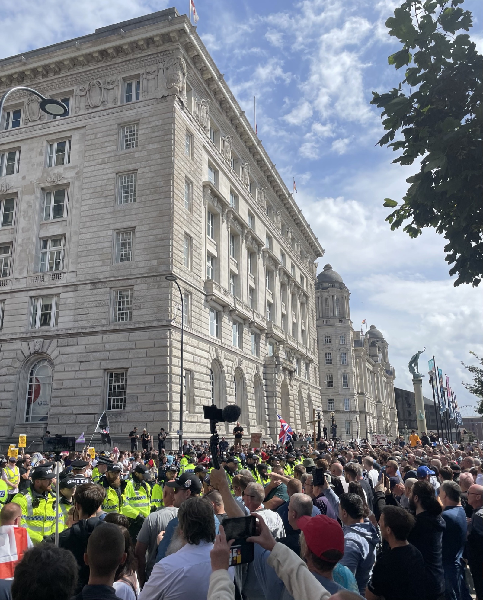 Una multitud, con alguna bandera británica, frente a un gran edificio de piedra, con una fila de policías con chaleco amarillo a la izquierda.