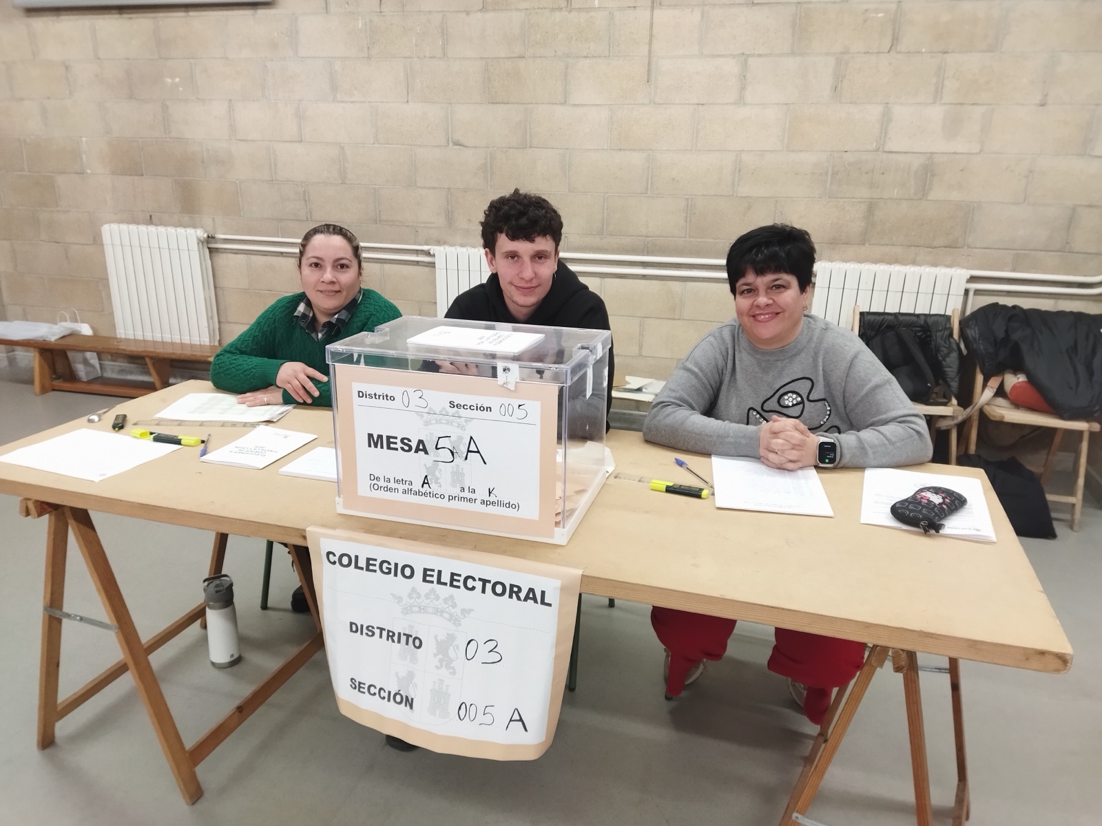 Tres personas sentadas tras una mesa con una urna transparente y el cartel «Mesa 5A», en un colegio electoral.