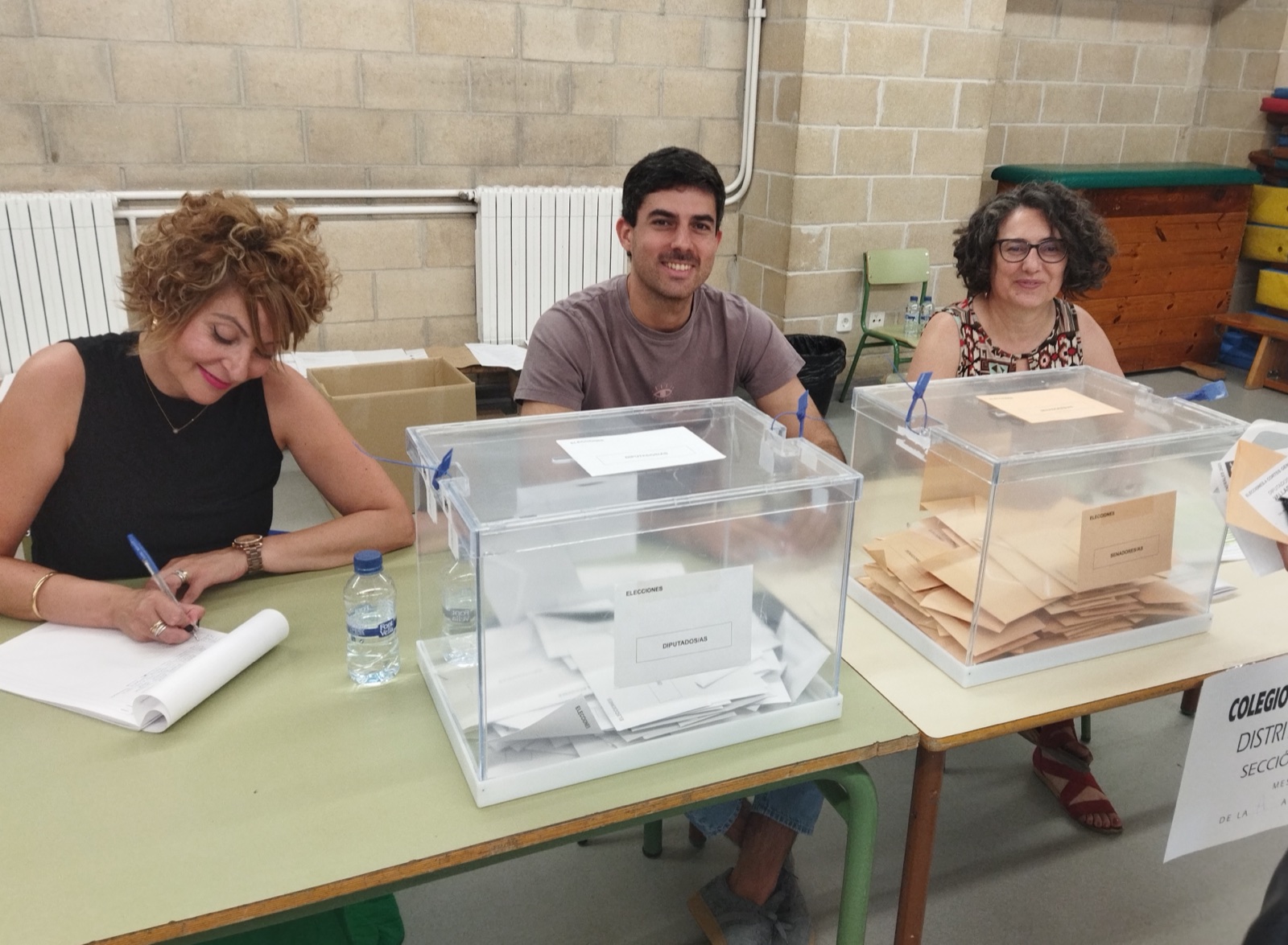 Tres miembros de una mesa electoral sentados tras dos urnas transparentes con sobres.