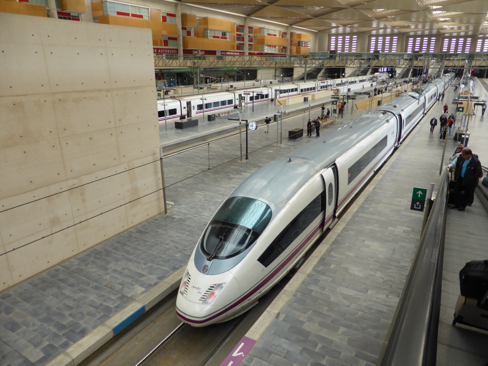 Un tren de alta velocidad blanco con el morro gris parado junto a un andén en una gran estación cubierta, con otro tren al fondo.