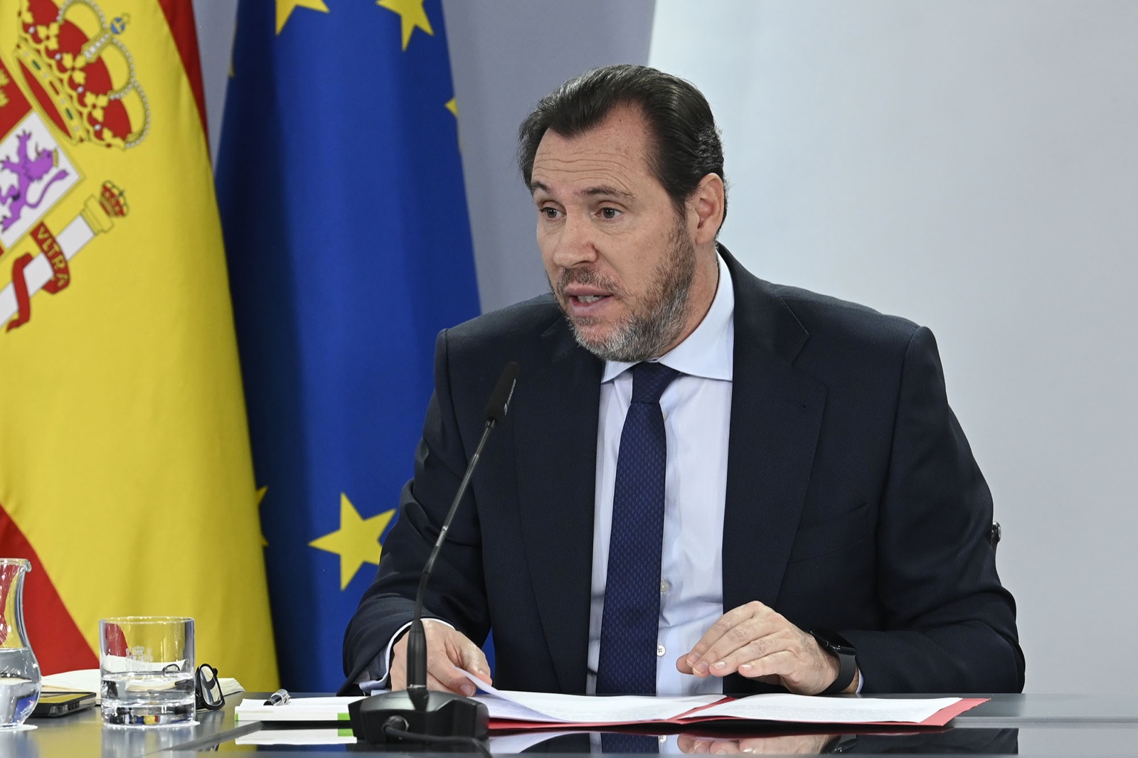 Un hombre con barba, traje oscuro y corbata azul habla ante un micrófono sentado a una mesa, con las banderas de España y de la Unión Europea detrás.