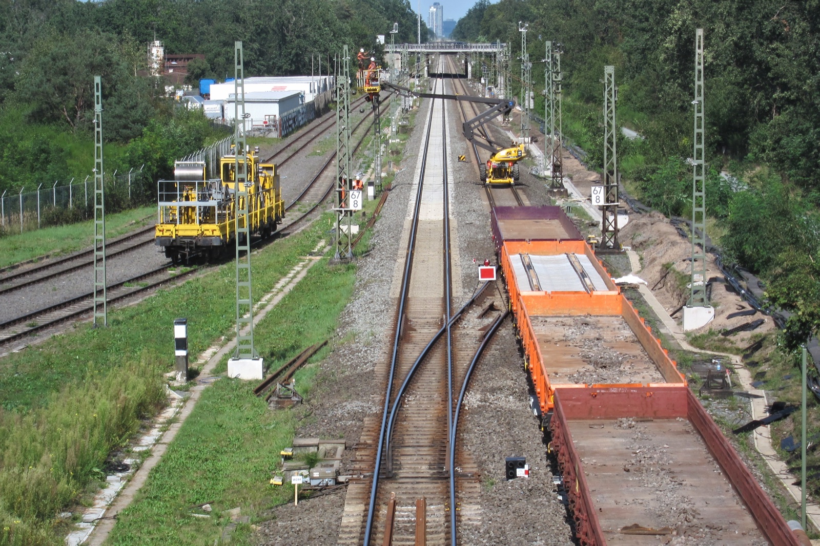 Vías de tren en obras con máquinas amarillas, vagones de carga naranjas con balasto y postes de catenaria, entre árboles.