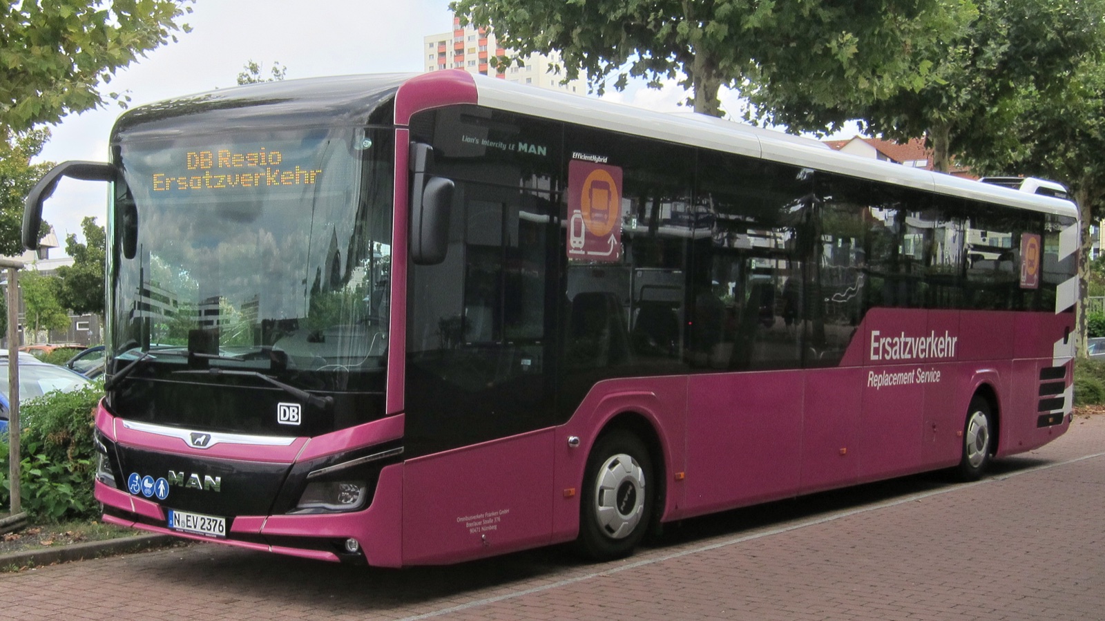 Autobús magenta y blanco con el rótulo «Ersatzverkehr» (servicio de sustitución) aparcado junto a una acera bajo unos árboles.