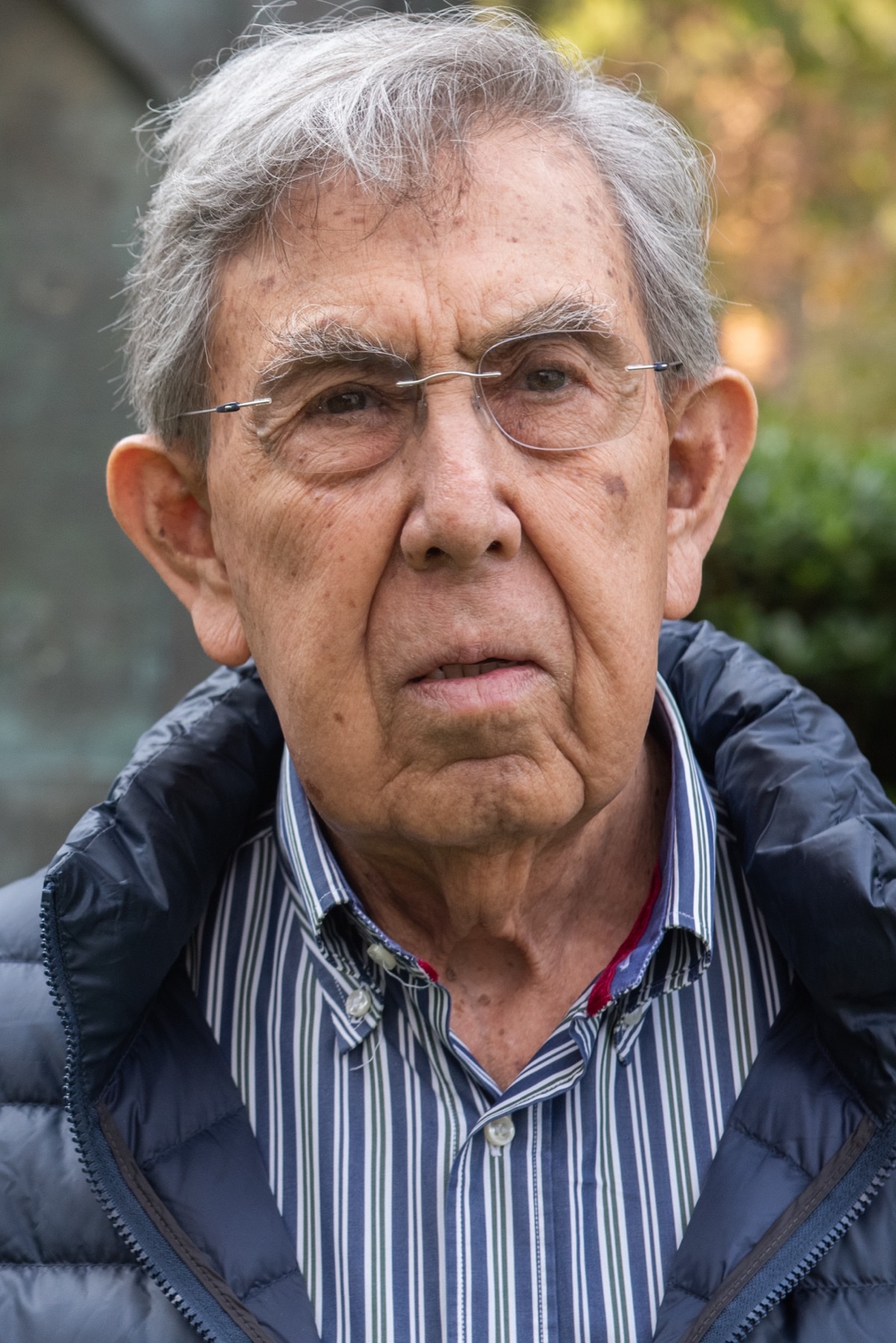 Retrato de Cuauhtémoc Cárdenas, de pelo canoso y con gafas sin montura, con camisa de rayas azules y un plumífero azul marino.