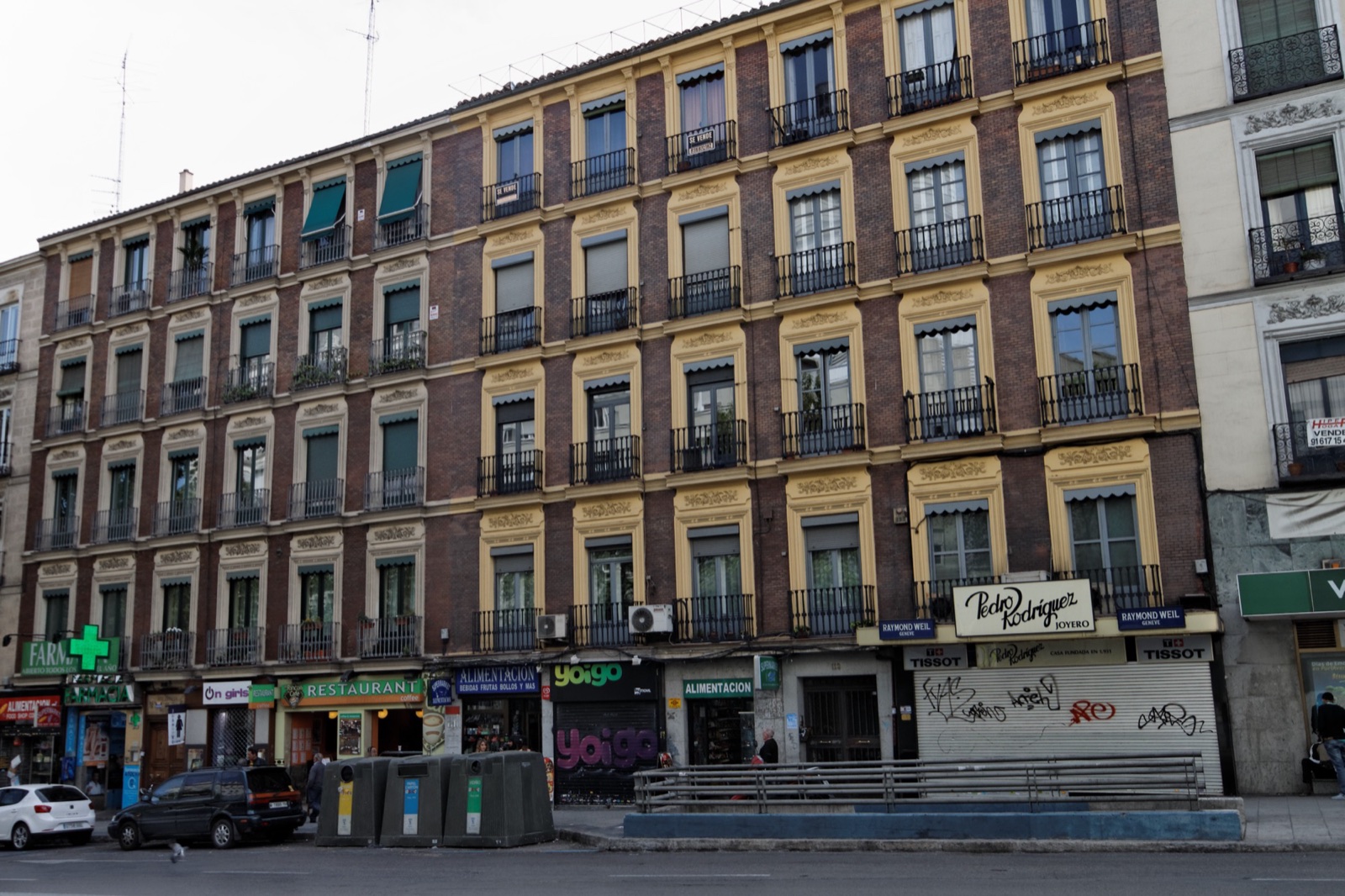 Fachada de edificios de viviendas con balcones de hierro, dos carteles de «Se vende» y comercios en los bajos.