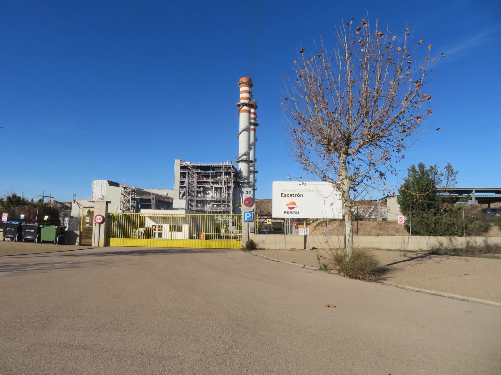 Entrada de la central de Escatrón, con sus dos chimeneas blancas y naranjas, las instalaciones de la planta y un cartel de Repsol, bajo un cielo azul.