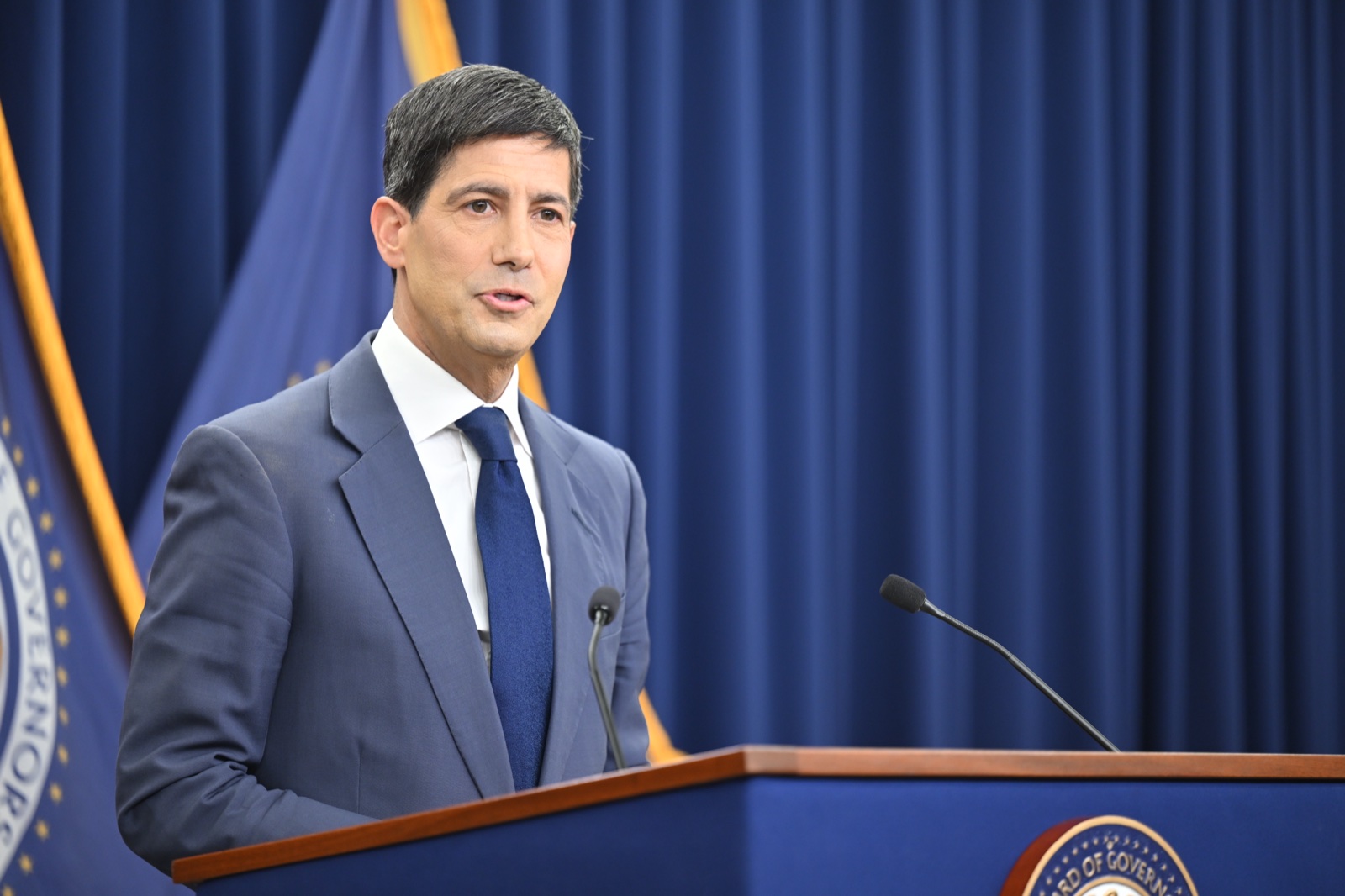 Kevin Warsh, con traje gris y corbata azul, habla en el atril de la Reserva Federal ante una cortina azul.
