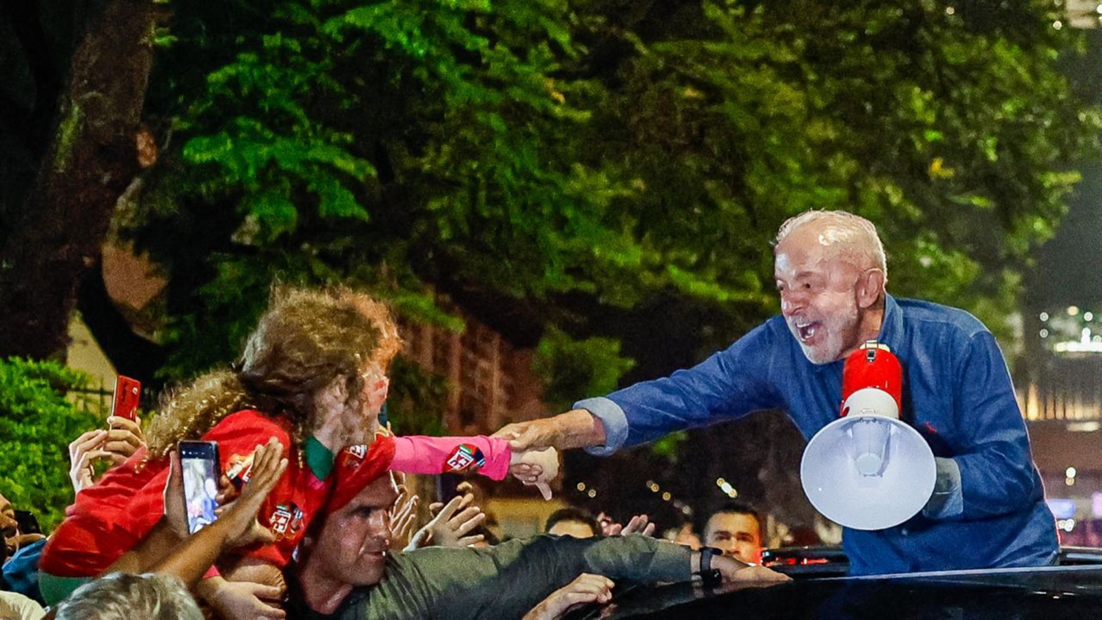 Lula, sonriente y con un megáfono en la mano, saluda desde un vehículo a una multitud de simpatizantes por la noche.