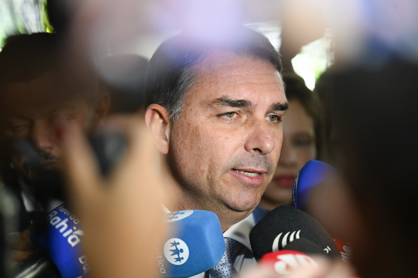 Flávio Bolsonaro, con camisa blanca y corbata, habla rodeado de micrófonos de varias emisoras.