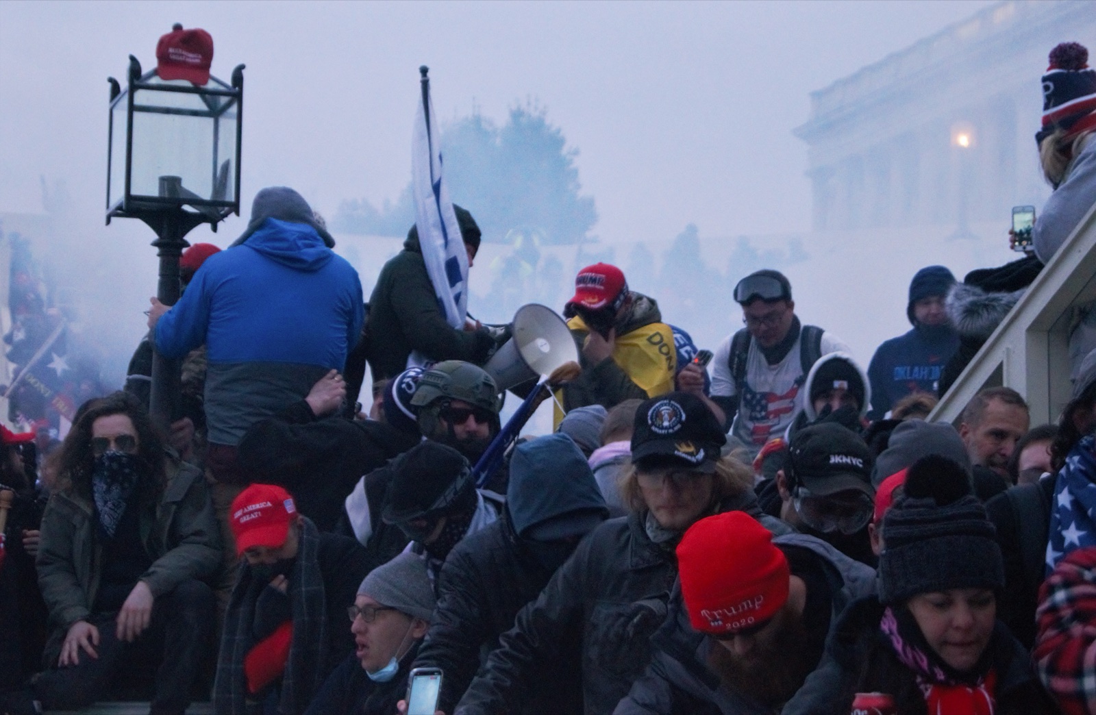 Una multitud con gorras rojas, banderas y un megáfono se agolpa en una escalinata envuelta en humo junto a la fachada del Capitolio.