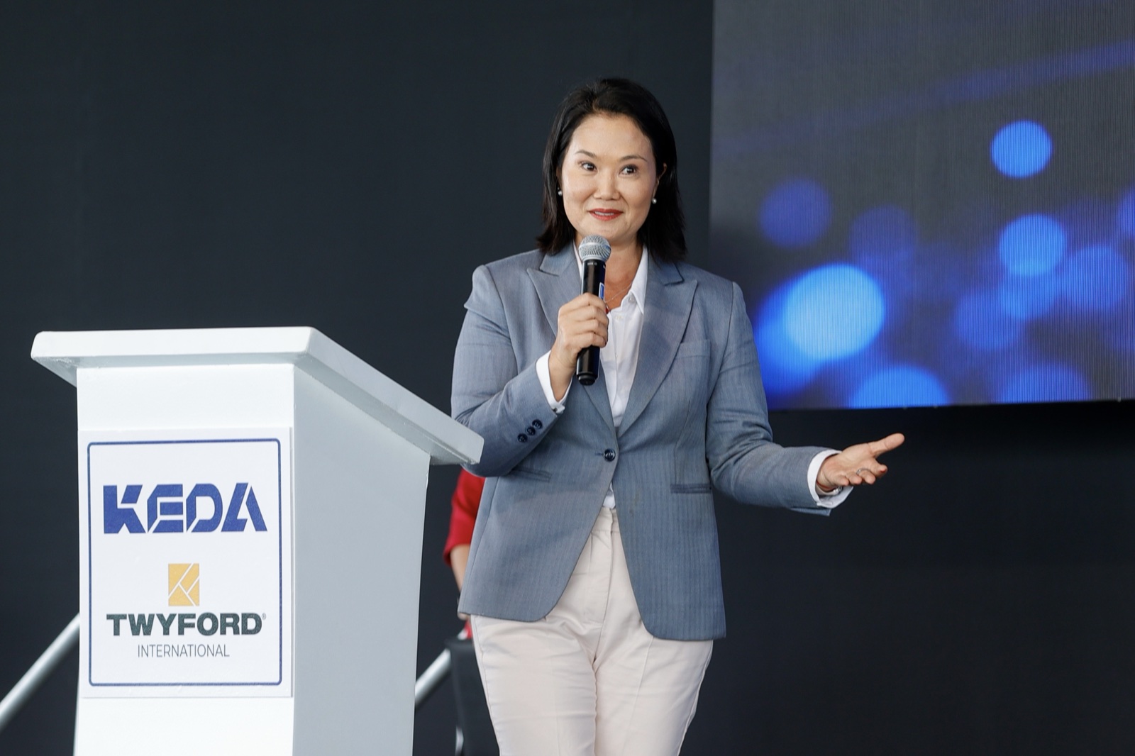Keiko Fujimori, con chaqueta gris y micrófono en la mano, habla en un escenario junto a un atril blanco.