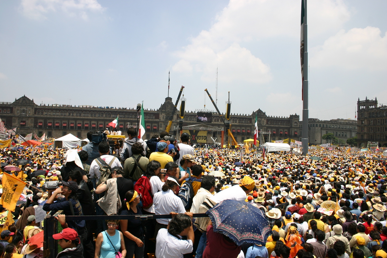 Una multitud con sombreros y banderas amarillas llena la plaza del Zócalo ante el Palacio Nacional, con un escenario al fondo.