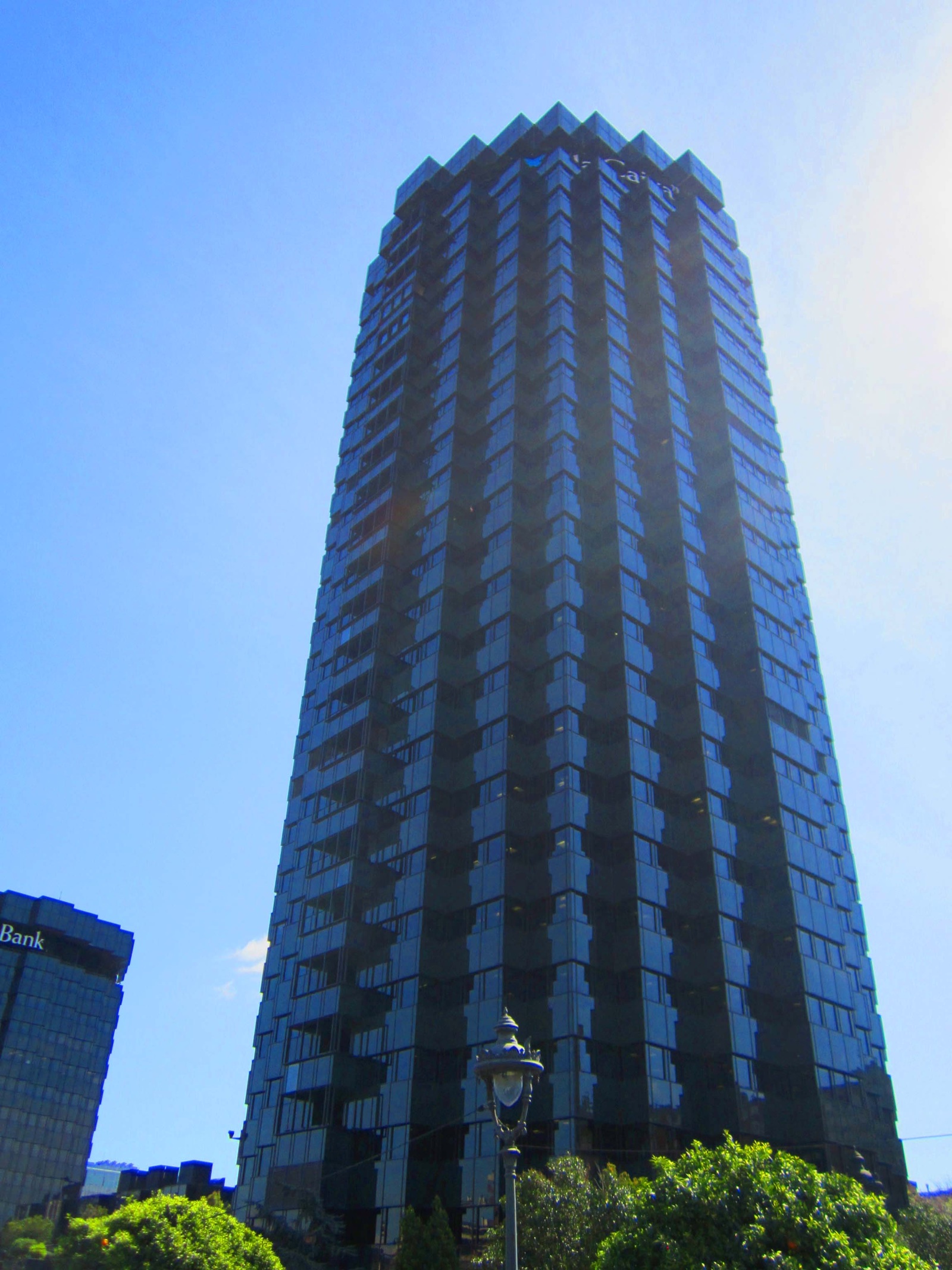 Torre de oficinas de cristal oscuro vista desde abajo contra un cielo azul, con el logotipo de la Caixa en lo alto y árboles y una farola en primer plano.