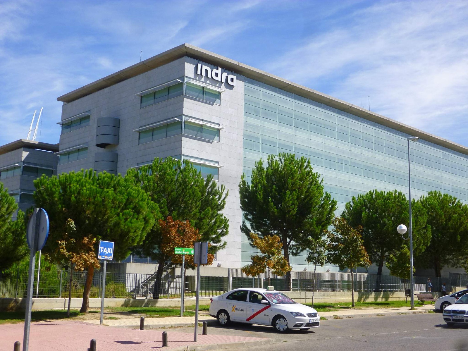 Edificio de oficinas de fachada gris y cristal con el rótulo «indra» en lo alto, con pinos delante y un taxi blanco aparcado en la calle.
