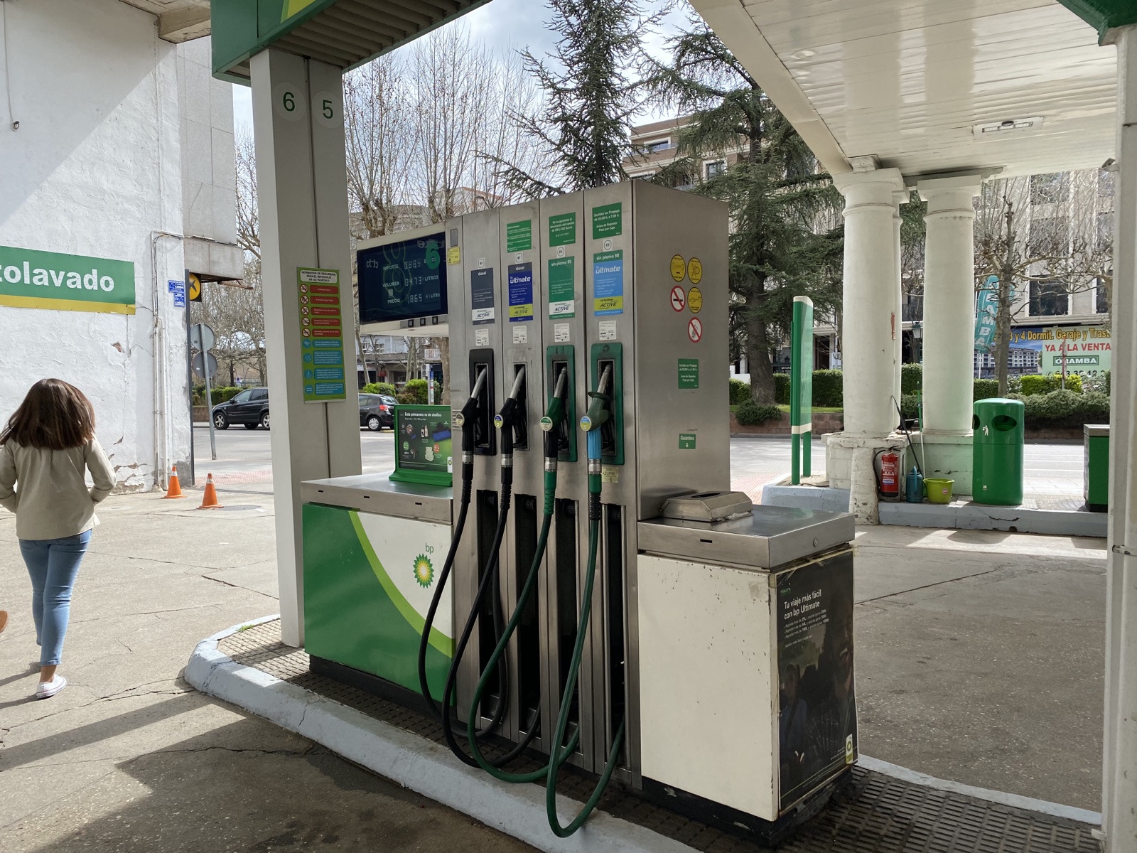 Surtidor de una gasolinera con cuatro mangueras, bajo la marquesina, con la pantalla del importe y los litros y una mujer caminando a la izquierda.