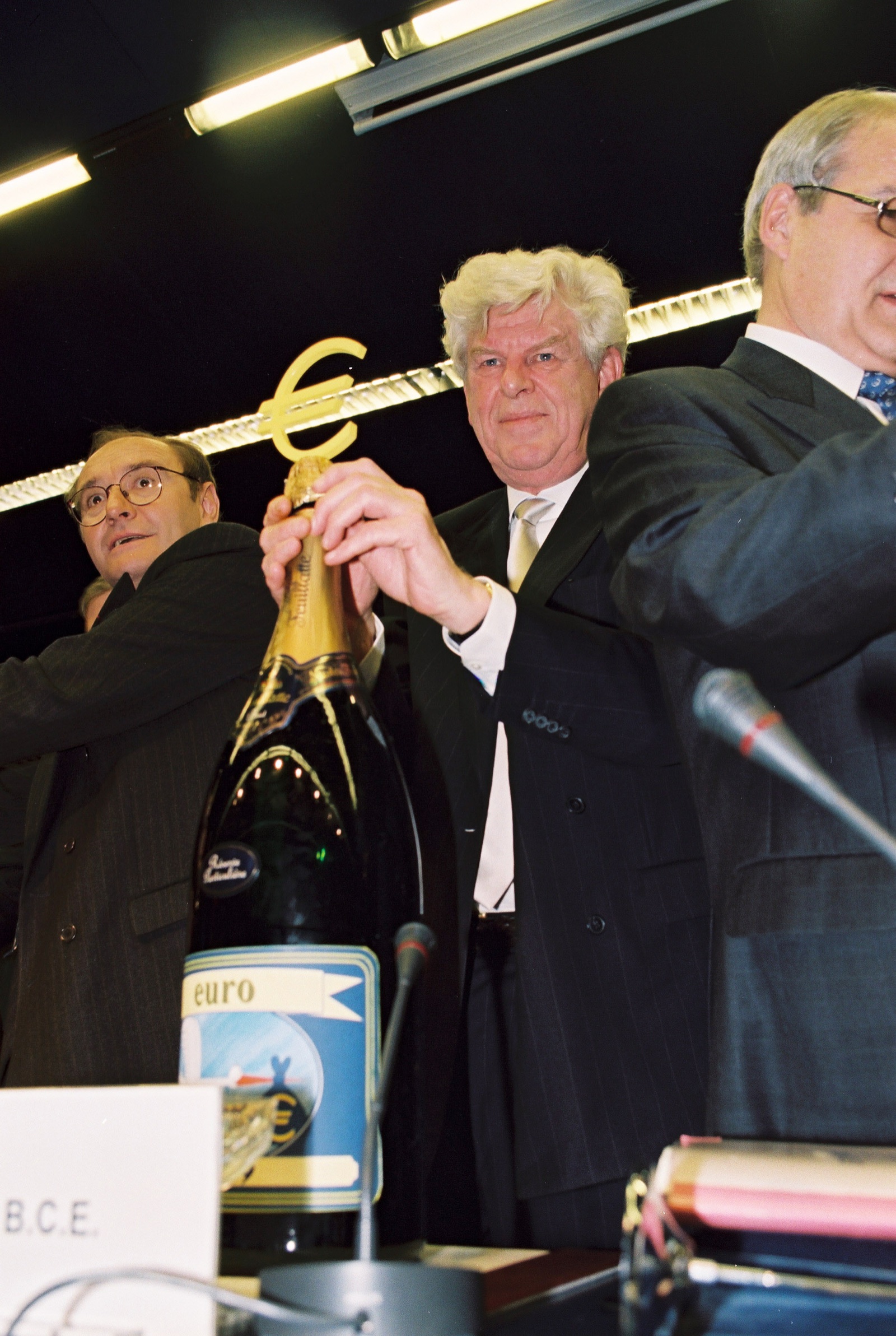 Un hombre de pelo blanco sostiene una botella grande de champán con un símbolo del euro dorado, rodeado de otros hombres con traje.