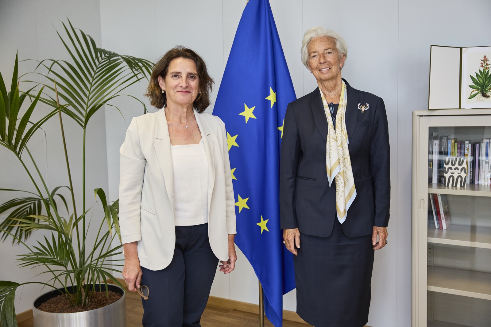 Dos mujeres posan de pie junto a una bandera de la Unión Europea en un despacho con plantas y una estantería.