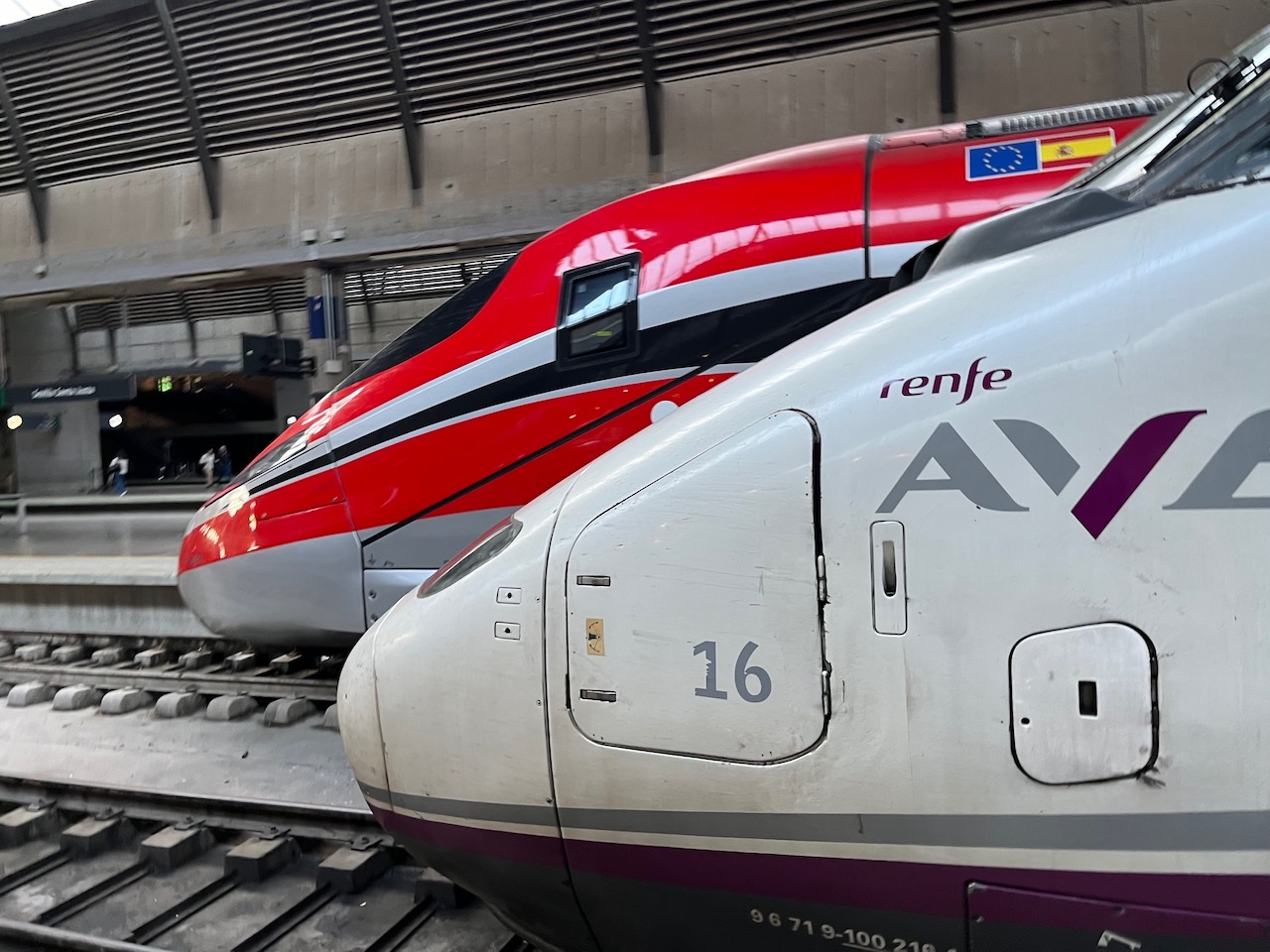 Morros de dos trenes de alta velocidad parados en andenes contiguos: en primer plano, uno blanco de Renfe con el rótulo AVE y, detrás, uno rojo de Iryo.