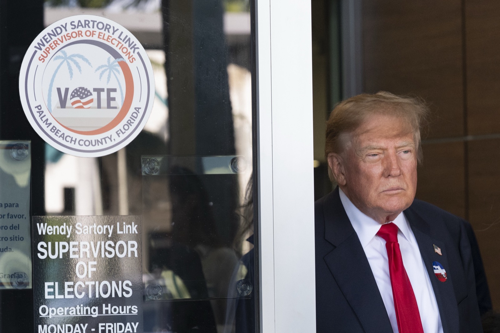 Donald Trump, con traje oscuro, corbata roja y una pegatina de voto en la solapa, a la puerta de una oficina electoral con el cartel «Vote».