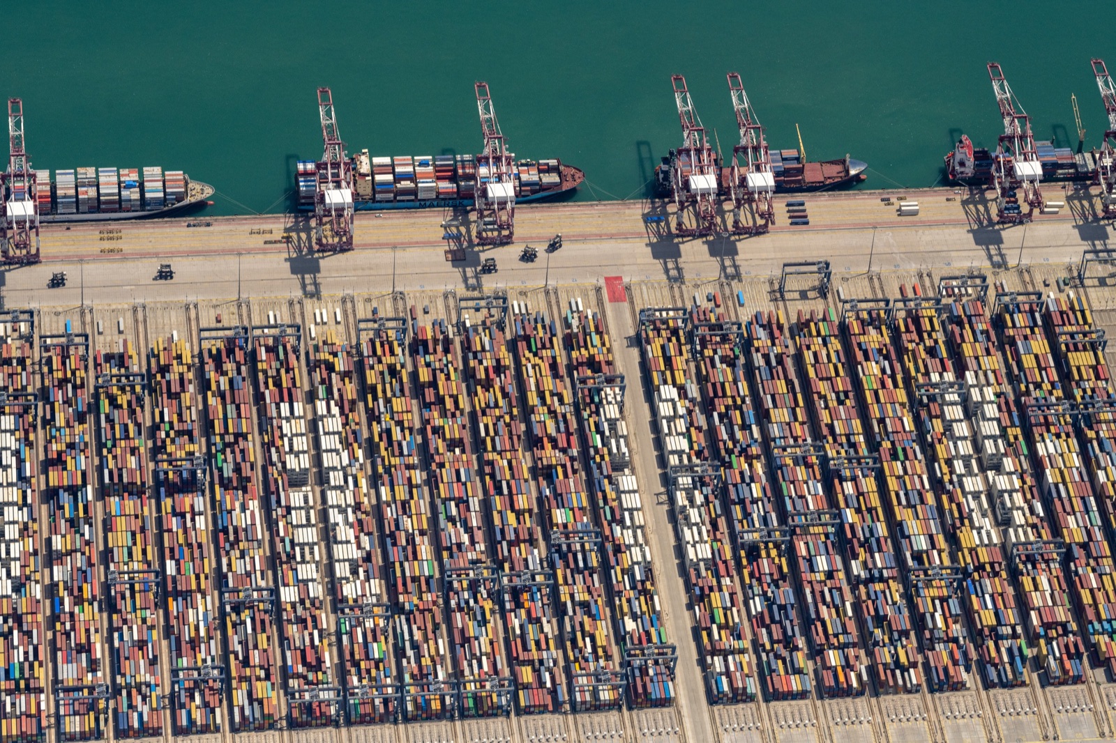 Vista aérea de una terminal de contenedores con filas de contenedores de colores, grúas en el muelle y varios buques atracados.