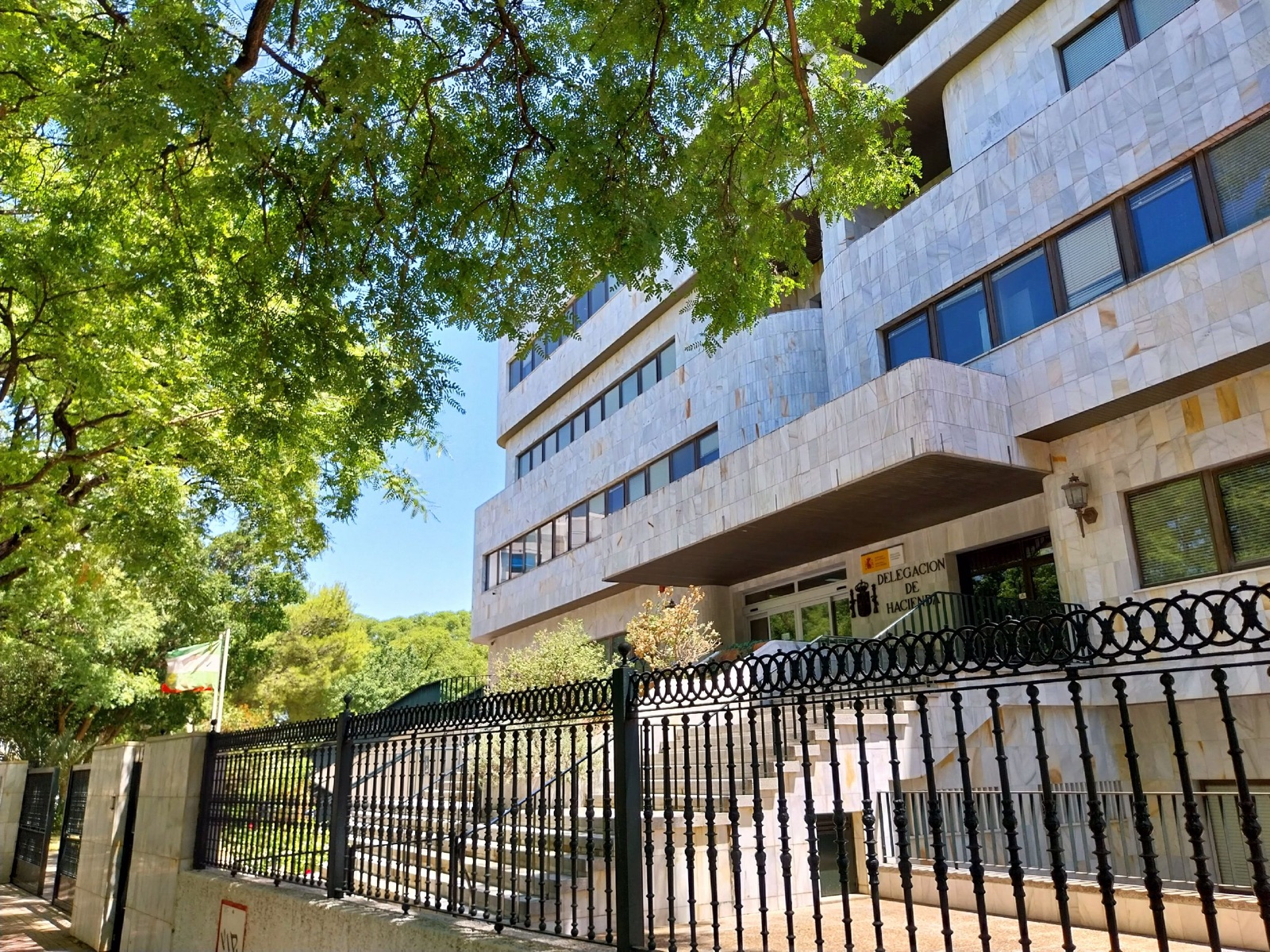 Edificio de oficinas revestido de piedra clara, con el rótulo «Delegación de Hacienda» junto a la entrada, tras una verja negra y bajo las ramas de un árbol.