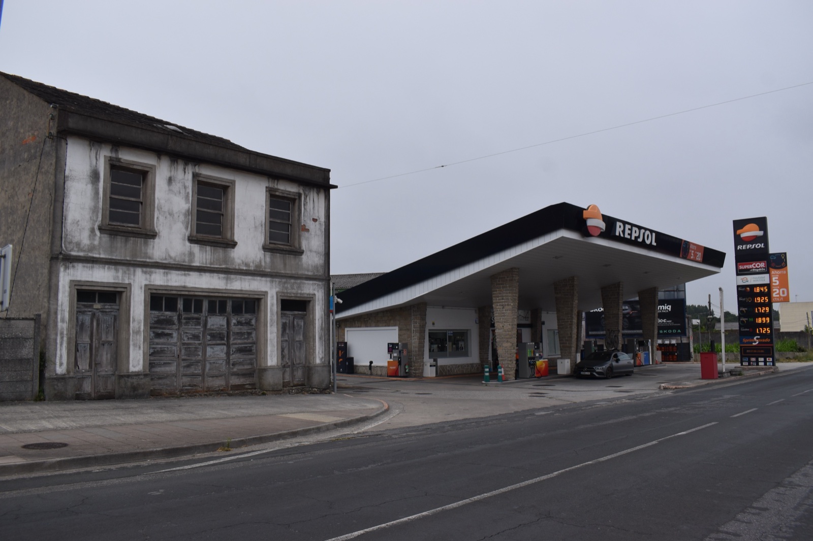 Gasolinera con la marquesina de Repsol junto a una carretera y un edificio antiguo, con un poste de precios a la derecha, bajo un cielo nublado.