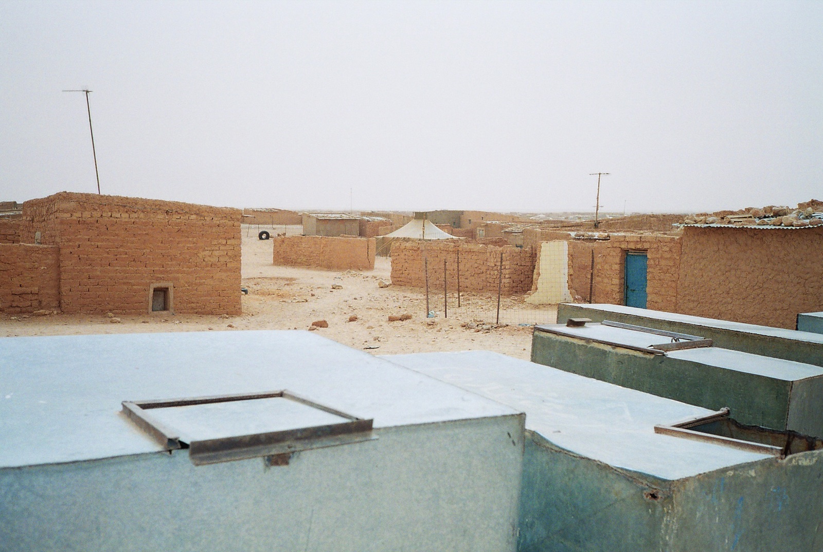 Casas bajas de adobe y depósitos de agua metálicos en primer plano, en un paisaje desértico bajo un cielo gris.