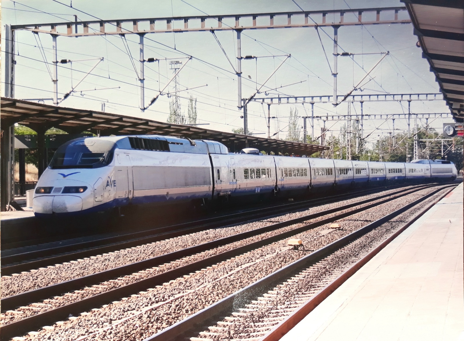 Un tren de alta velocidad blanco con el rótulo AVE detenido junto a un andén, bajo la catenaria, con varias vías en primer plano.
