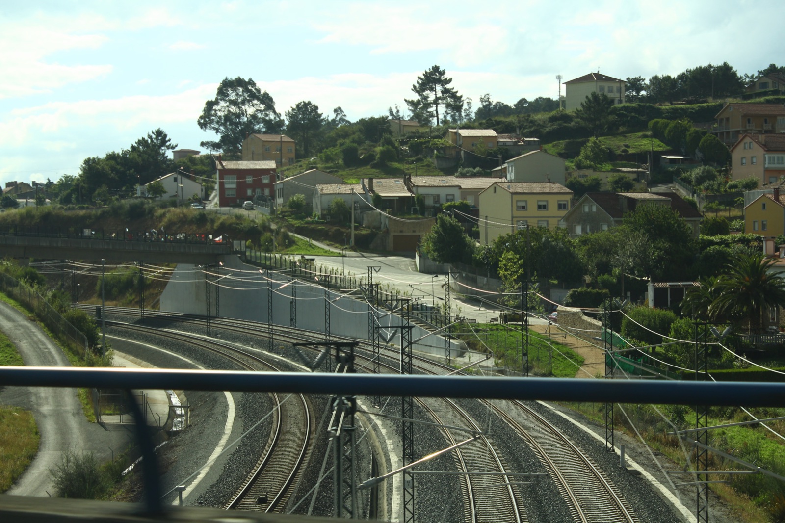Vías de tren que trazan una curva bajo la catenaria, vistas desde un puente, con casas en una ladera al fondo.