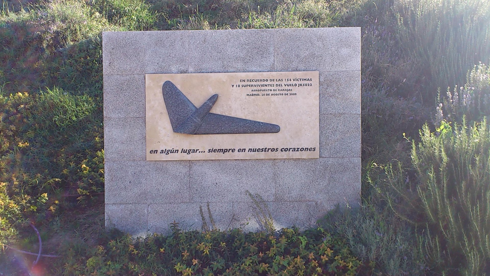 Monolito de piedra con una placa clara que lleva la silueta de un avión y el texto «en algún lugar... siempre en nuestros corazones», entre arbustos.
