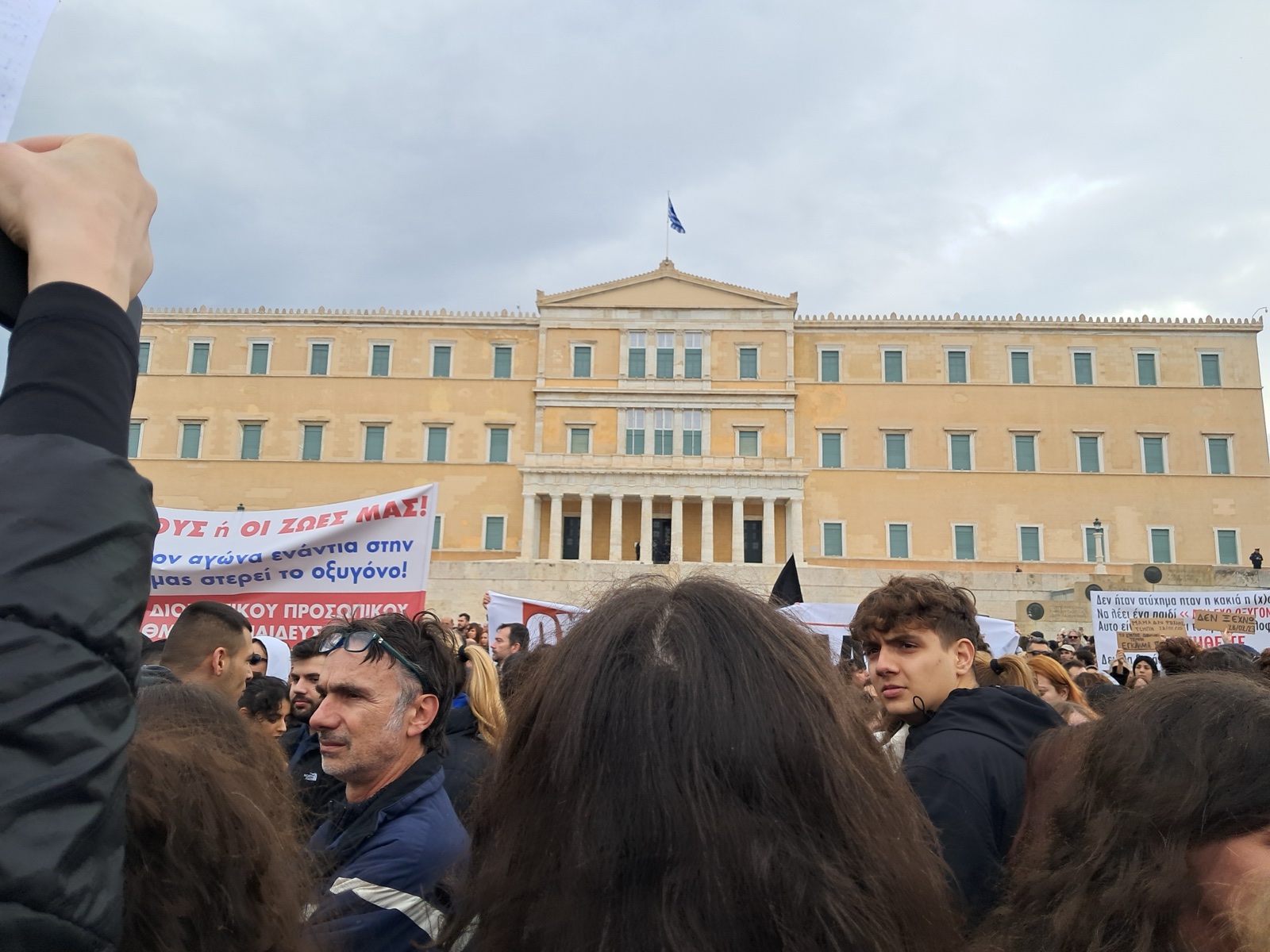 Una multitud con pancartas ante un gran edificio neoclásico de color ocre con la bandera de Grecia en lo alto.
