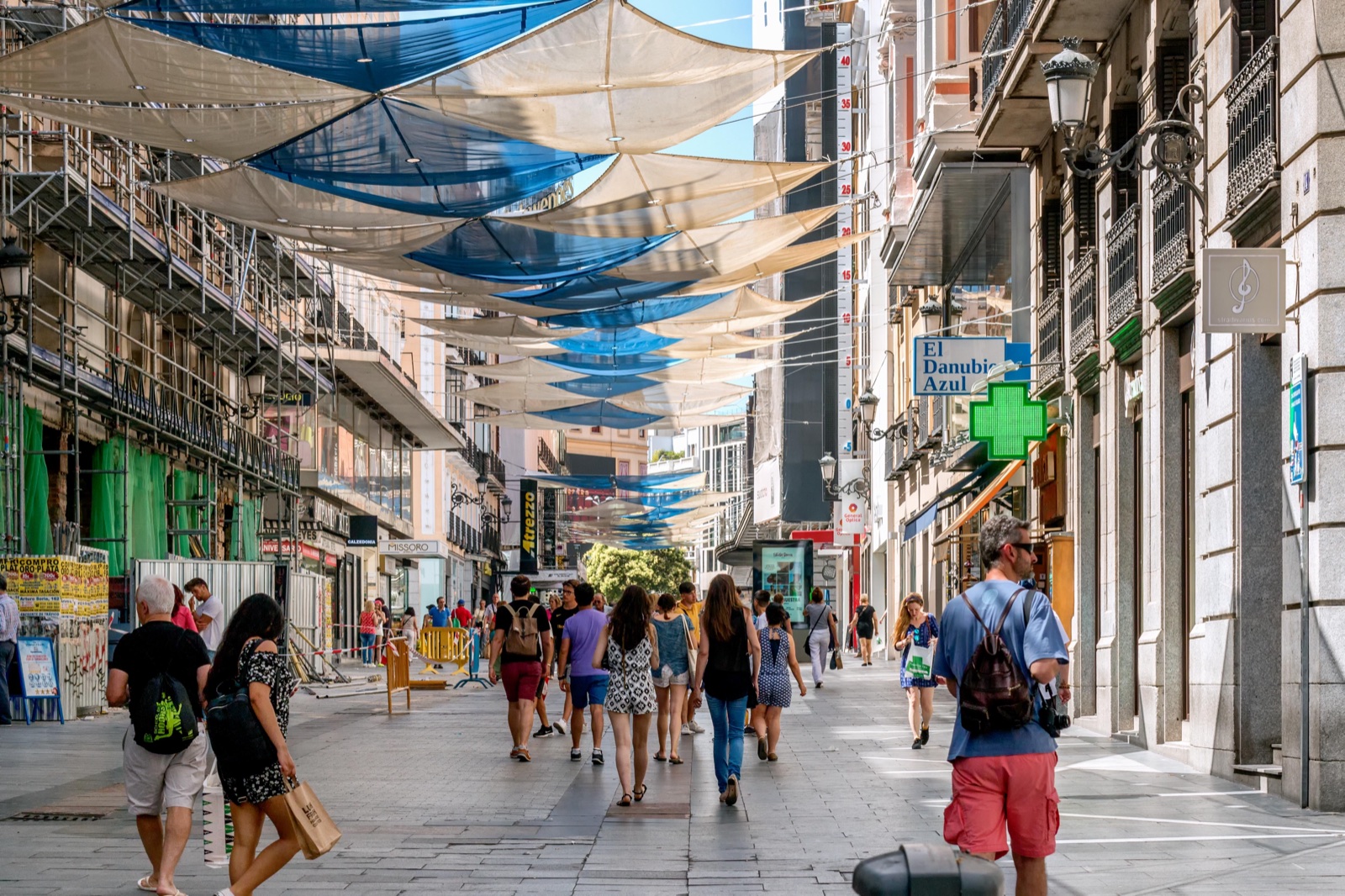 Calle peatonal comercial con toldos de tela tendidos entre los edificios y gente paseando, algunos con bolsas de compras, junto a escaparates y la cruz verde de una farmacia.